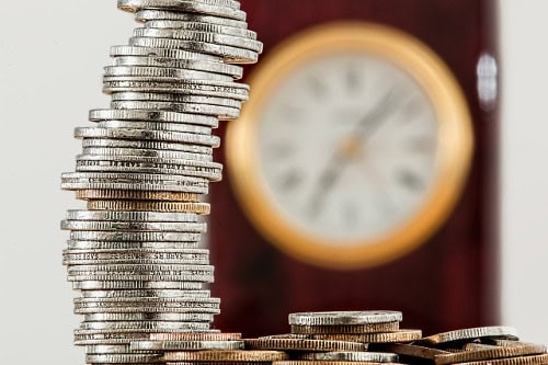 Coins stacked in a tall, curved formation with a blurred clock behind.